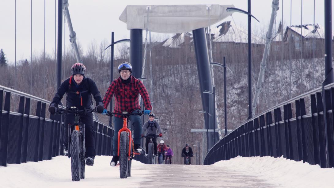 Two men in winter parkas and helmets lead a group of fat bike cyclists over a bridge.